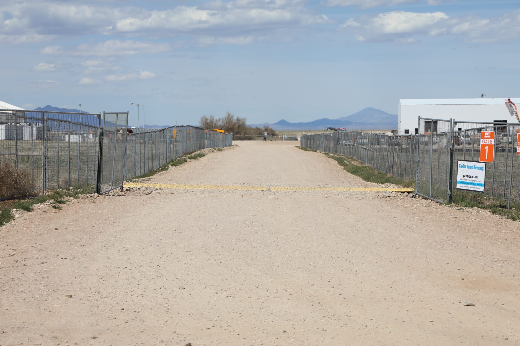 Chain link temporary fence lining both sides of a gravel haul road at the American Pacific plant west of Cedar City, with a Cedar Temp Fencing sign on the gate